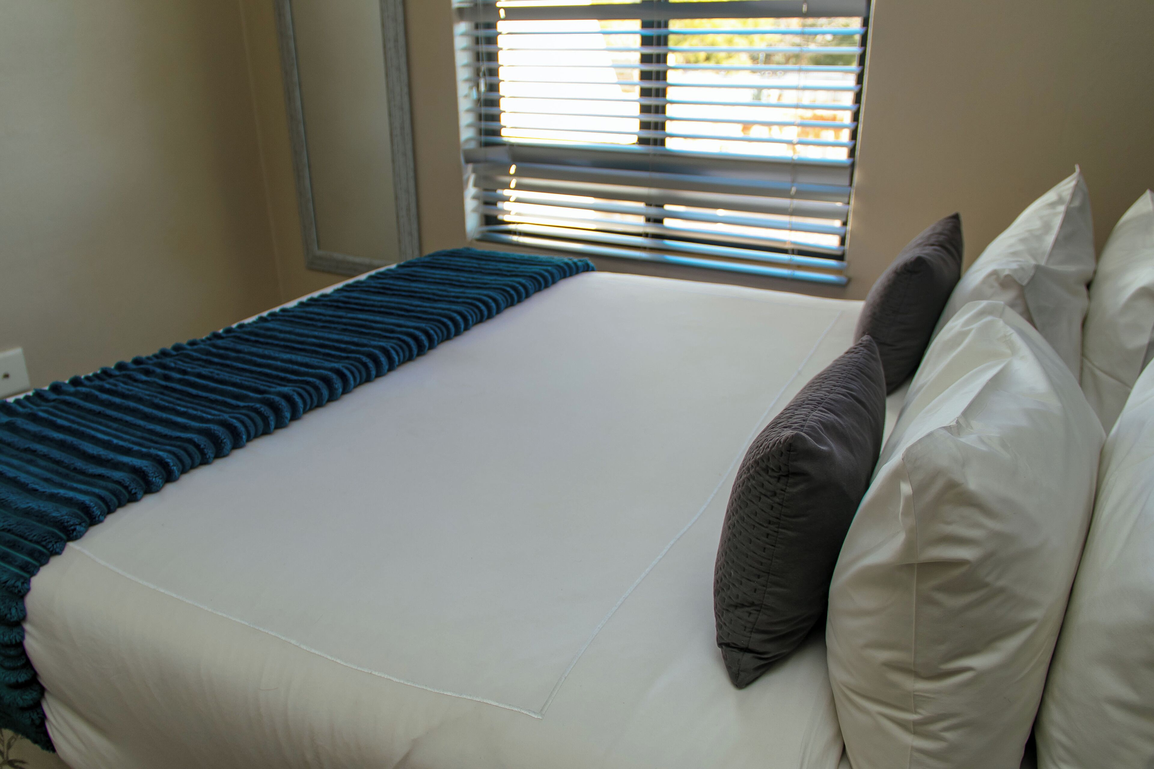 Una cama bien hecha con un corredor azul a rayas, almohadas blancas y una ventana con persianas.