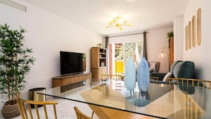 Ein modernes Wohnzimmer mit einem Glastisch, Holzsesseln, einem Fernseher und einer großen Topfpflanze.