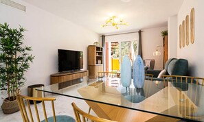 Ein modernes Wohnzimmer mit einem Glastisch, Holzsesseln, einem Fernseher und einer großen Topfpflanze.