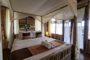 A large bed with a canopy, a bedside table with a lamp, and a view of the outside through a window. - Room-charming Deluxe Room Upper Floor at (Ubud)