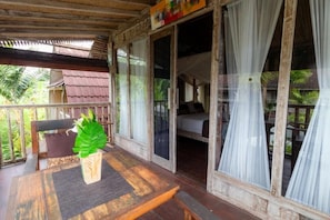 A wooden veranda with a table, a chair, and a bed visible through the glass doors. - Room-charming Deluxe Room Upper Floor at (Ubud)