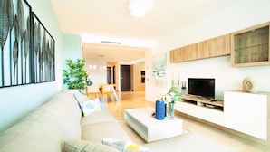 A modern living room with a sofa, coffee table, and TV stand. There is a dining area in the background with a table and chairs. - South Forest 9-1 (Ojén)