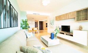 Ein modernes Wohnzimmer mit Sofa, Couchtisch und TV-Schrank. Im Hintergrund ist eine Essecke mit Tisch und Stühlen zu sehen.