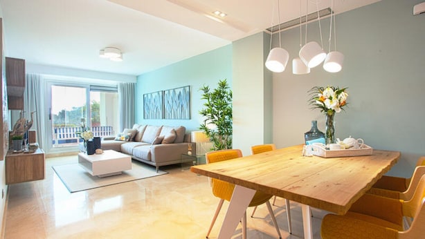 A modern living room with a sofa, coffee table, and dining table with chairs. There is a large window providing natural light and a view of the outdoors. - South Forest 9-1 (Ojén)
