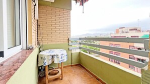 Un piccolo balcone con uno sgabello di legno, un tavolo con tovaglia a fiori e vista sui palazzi sullo sfondo.