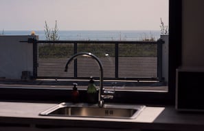 Une cuisine équipée d’un évier en acier inoxydable, d’un robinet et offrant une vue sur la mer à travers une clôture.