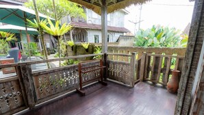 A wooden balcony with a thatched roof, surrounded by lush greenery and traditional buildings. - Room-deluxe Double or Twin Room (Ubud)