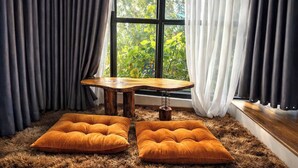 A room with a wooden table and two orange cushions on a furry rug, a window with curtains, and a view of greenery outside. - Gió Homestay Villa (Xuan Huong - Da Lat)