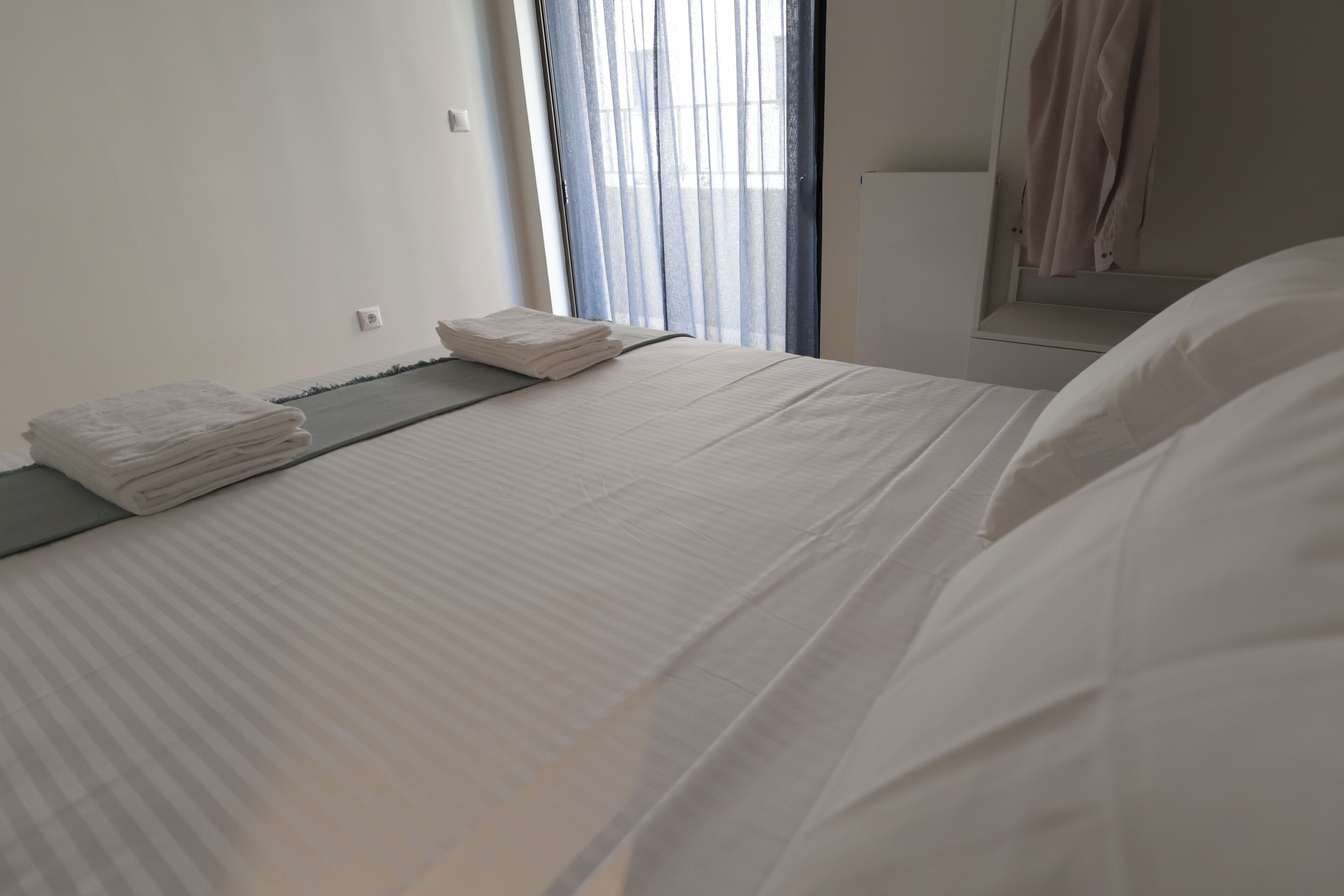 A neatly made bed with folded towels, a window with sheer curtains, and a white wall with a light switch.