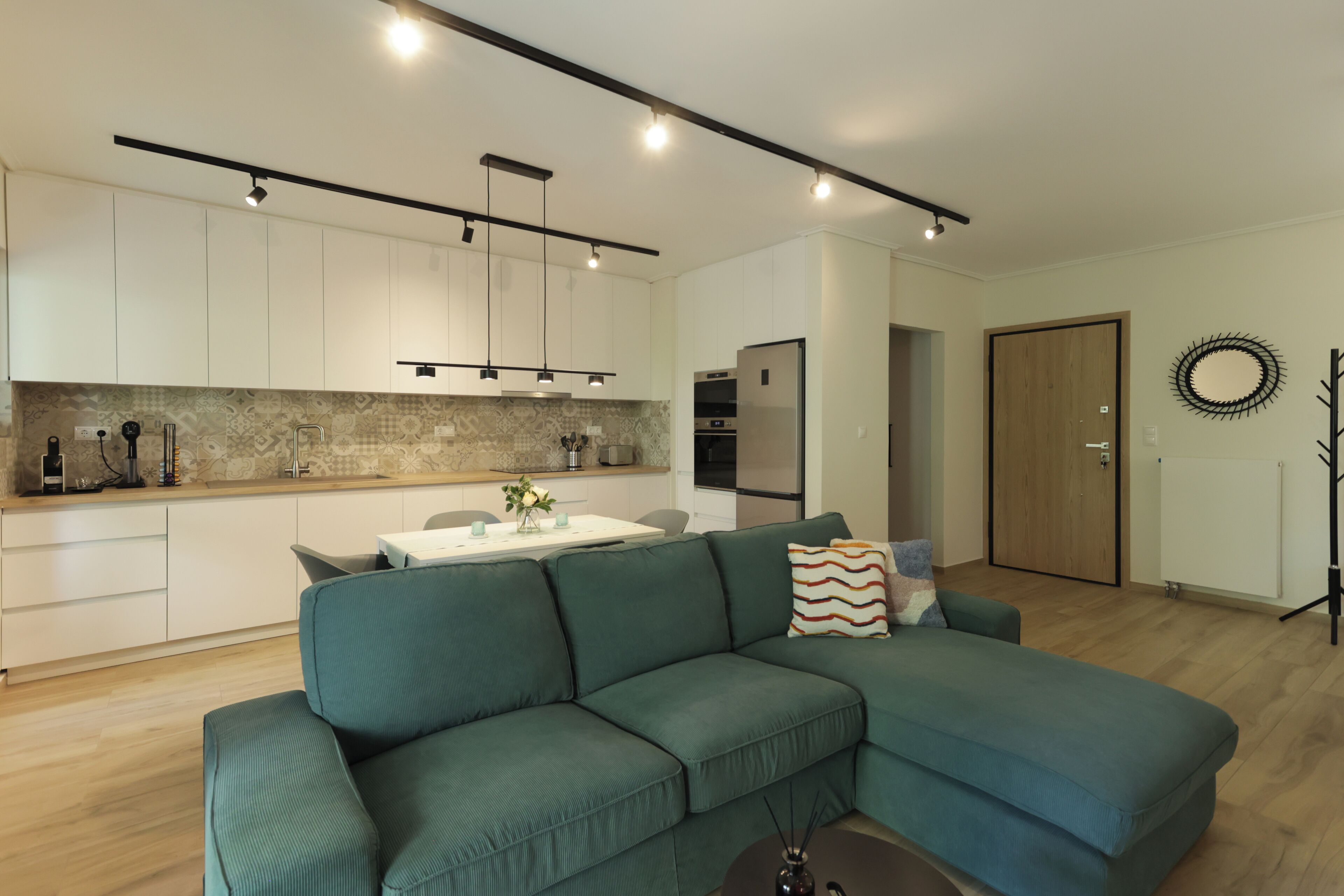 A modern living room with a green sectional sofa, a dining table, and a kitchen area with white cabinets and a backsplash.