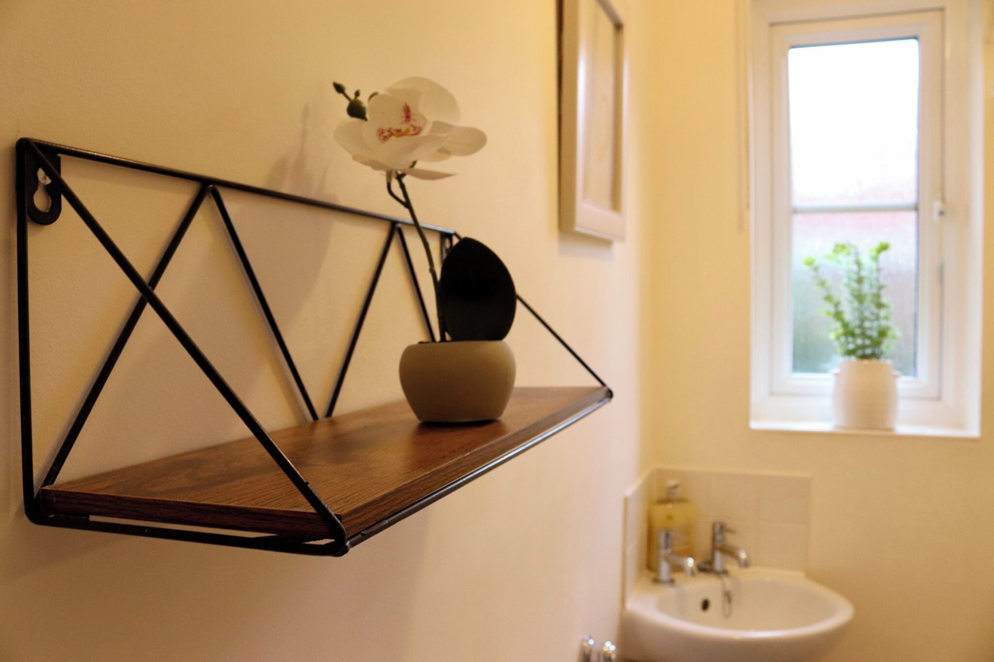 A bathroom with a wooden shelf holding a vase and an orchid, a white sink, and a window with a view of greenery.