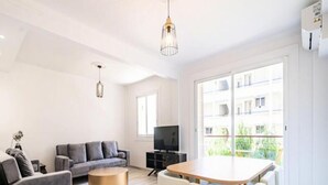 Ein modernes Wohnzimmer mit grauem Sofa, einem runden Couchtisch, einem Esstisch mit Stühlen, einem Flachbildfernseher und großen Fenstern mit Blick auf ein Gebäude und Grünflächen.