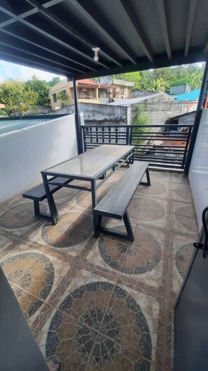 Eine überdachte Terrasse im Freien mit Tisch und Bänken, einem Fußboden aus gemusterten Fliesen und Blick auf die umliegenden Gebäude.
