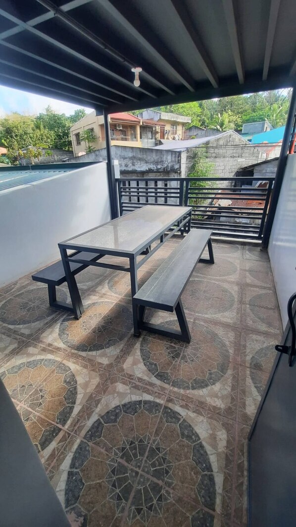 Une terrasse extérieure couverte, dotée d’une table et de bancs, de carreaux de sol à motifs et offrant une vue sur les bâtiments environnants.