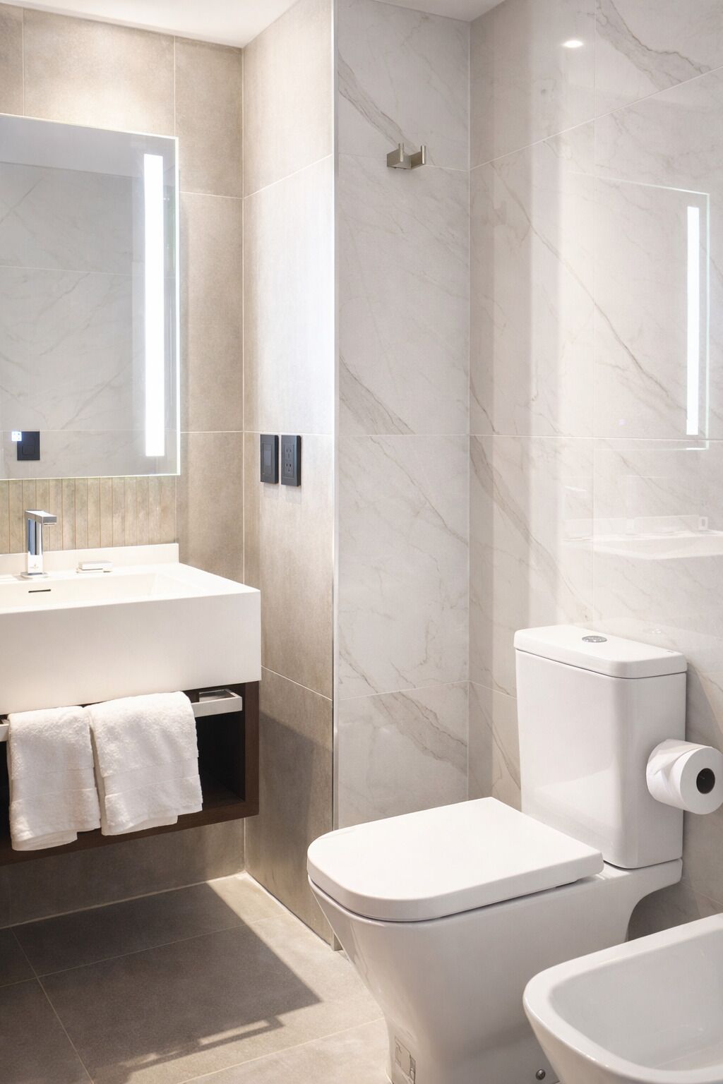 A modern bathroom with a marble wall, a white sink, a toilet, and a mirror.
