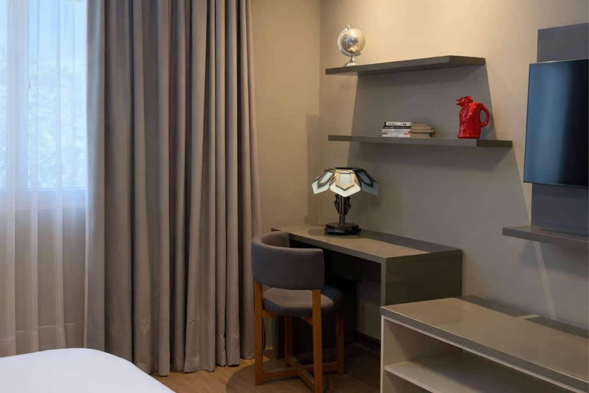 Et hotelværelse med seng, skrivebord med lampe, stol, fjernsyn og en reol med globus og bøger.
