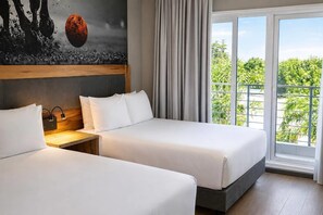 A hotel room with two beds, a wooden headboard, a nightstand, a wall-mounted lamp, and a large window with a view of greenery.