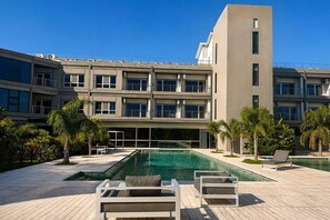 A modern building with a swimming pool, lounge chairs, and palm trees.
