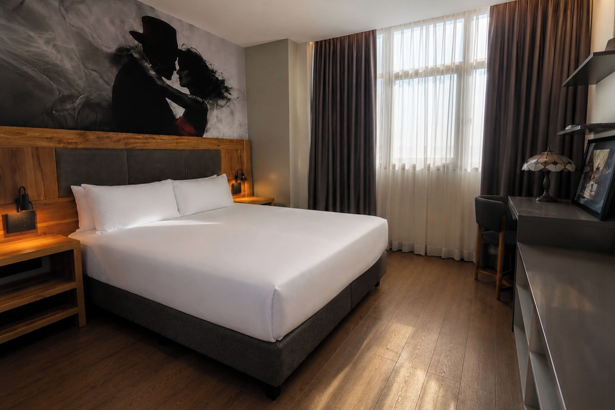 Una habitación de hotel con una cama grande, cabecera de madera, una mesita de noche, un escritorio con lámpara y un ventanal con cortinas.
