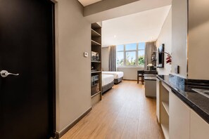 A modern hotel room with a bed, a desk, a kitchenette, and a large window.