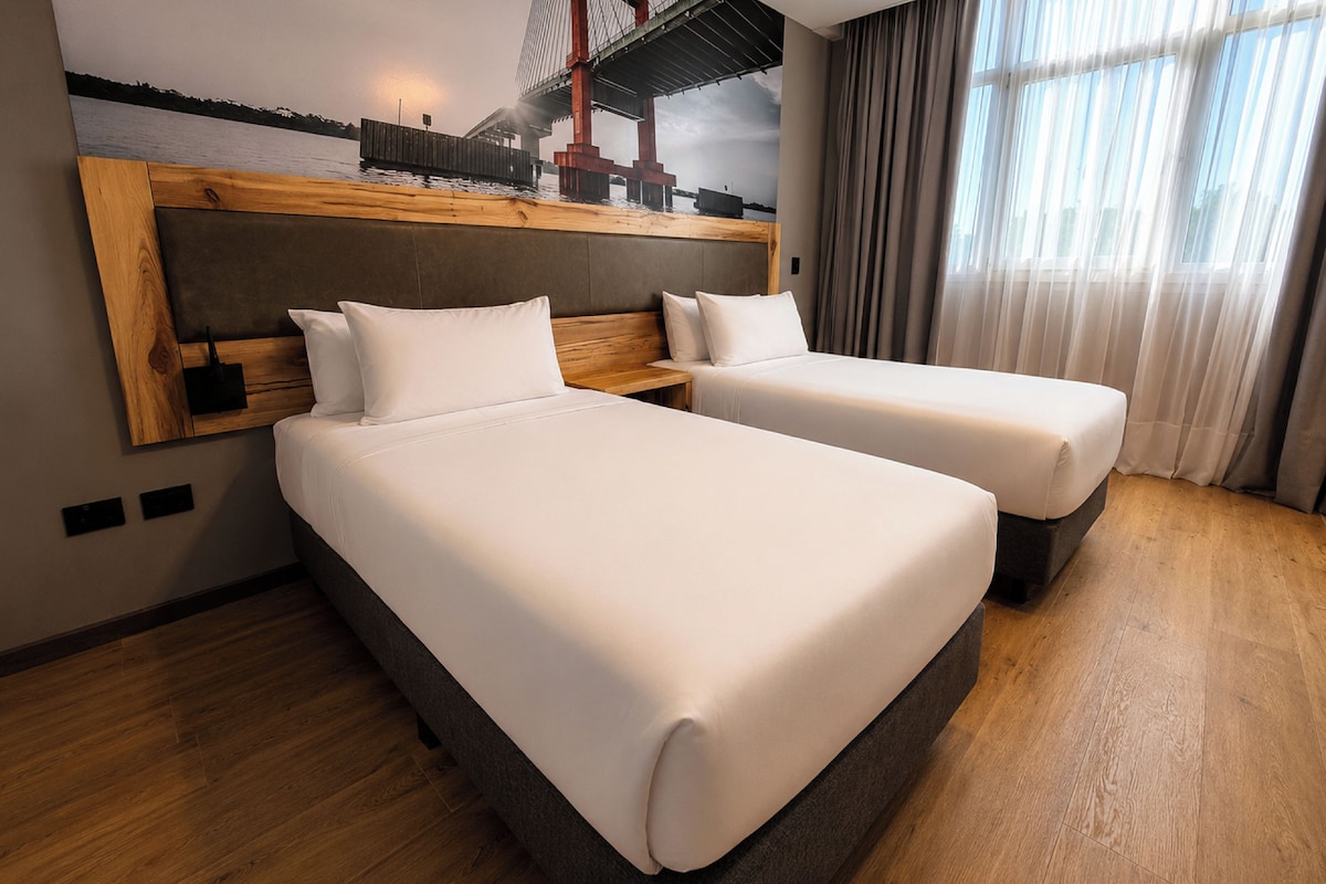 Habitación de hotel con dos camas, cabecera de madera, un ventanal con cortinas y un cuadro en la pared.