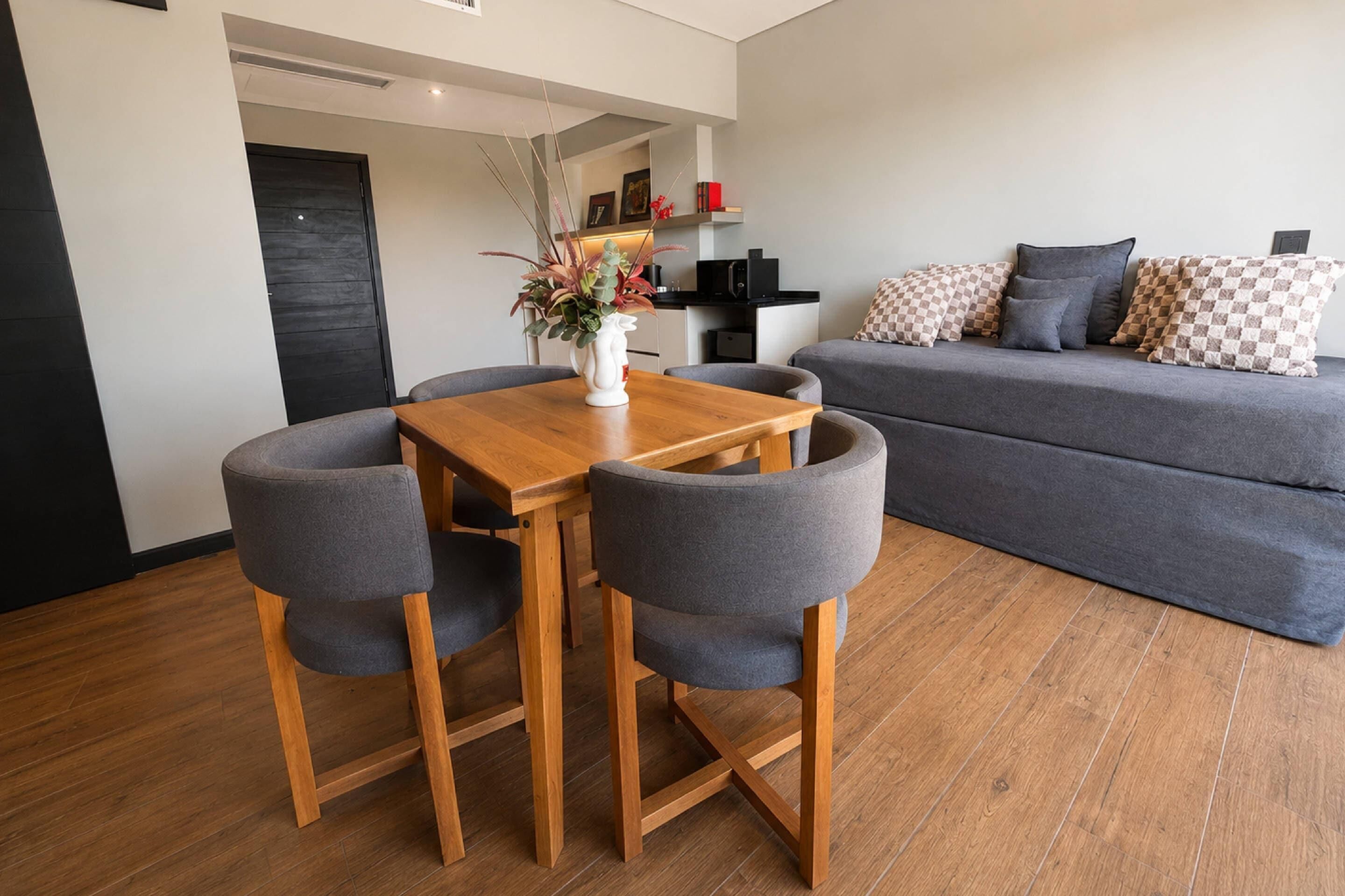 Un espace de vie moderne comprenant une table à manger, quatre chaises grises, un canapé gris et un coin cuisine à l’arrière-plan.