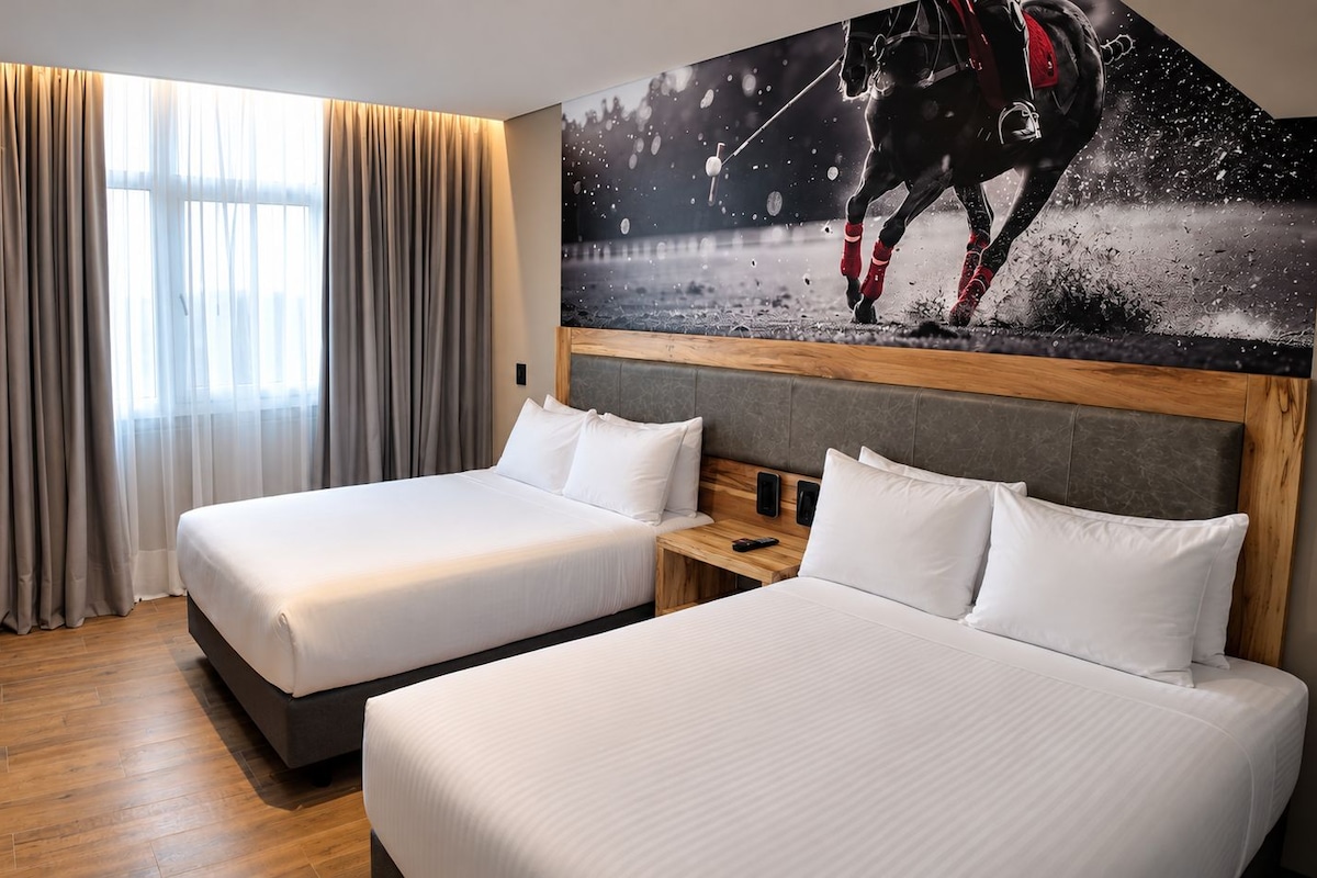 Habitación de hotel con dos camas, un ventanal con cortinas, un televisor montado y una obra de arte en la pared que muestra un caballo en movimiento.