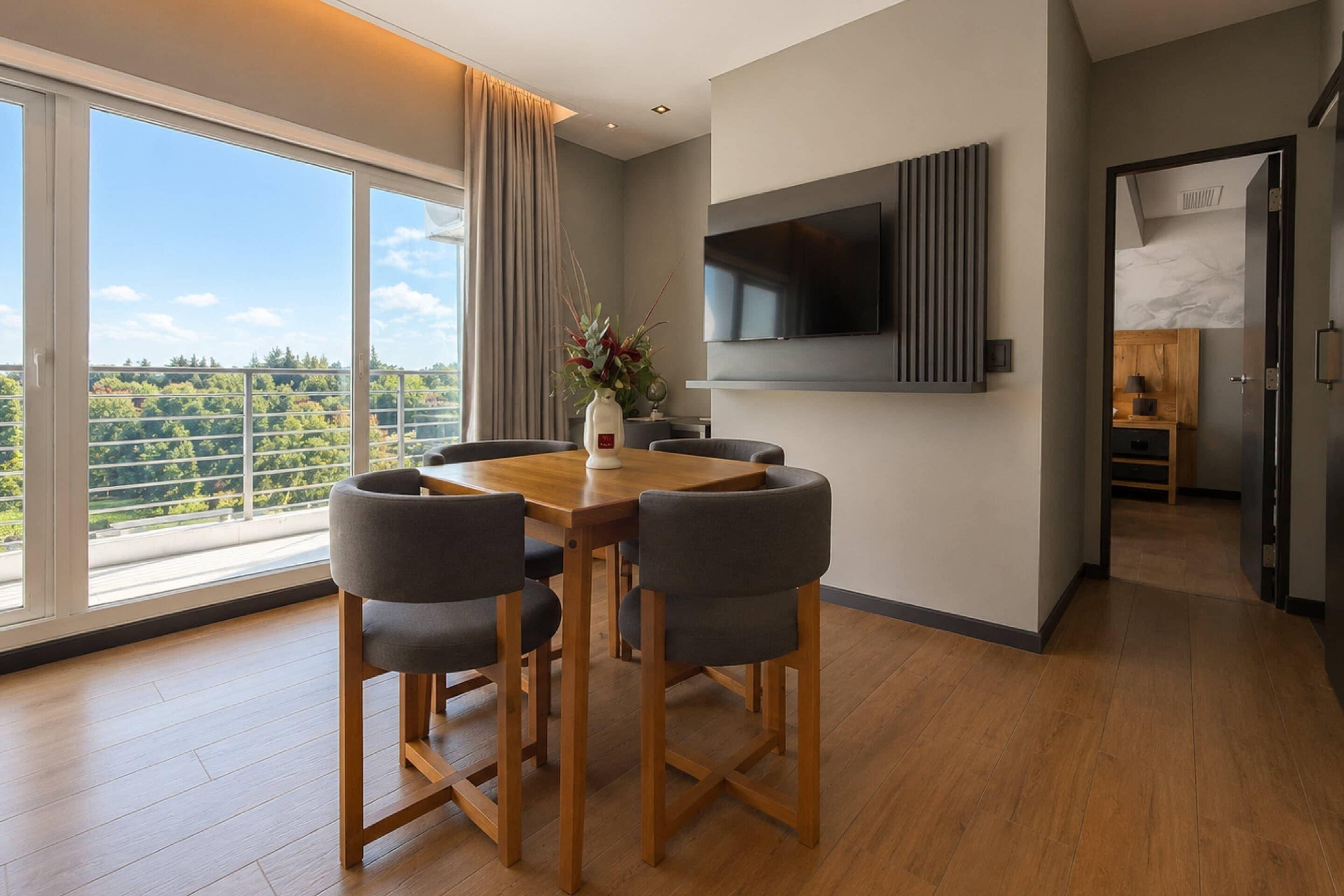 Un comedor moderno con una mesa y sillas de madera, un televisor montado en la pared y vistas a zonas verdes a través de amplias ventanas.