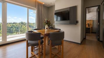 Un comedor moderno con una mesa y sillas de madera, un televisor montado en la pared y vistas a zonas verdes a través de amplias ventanas.
