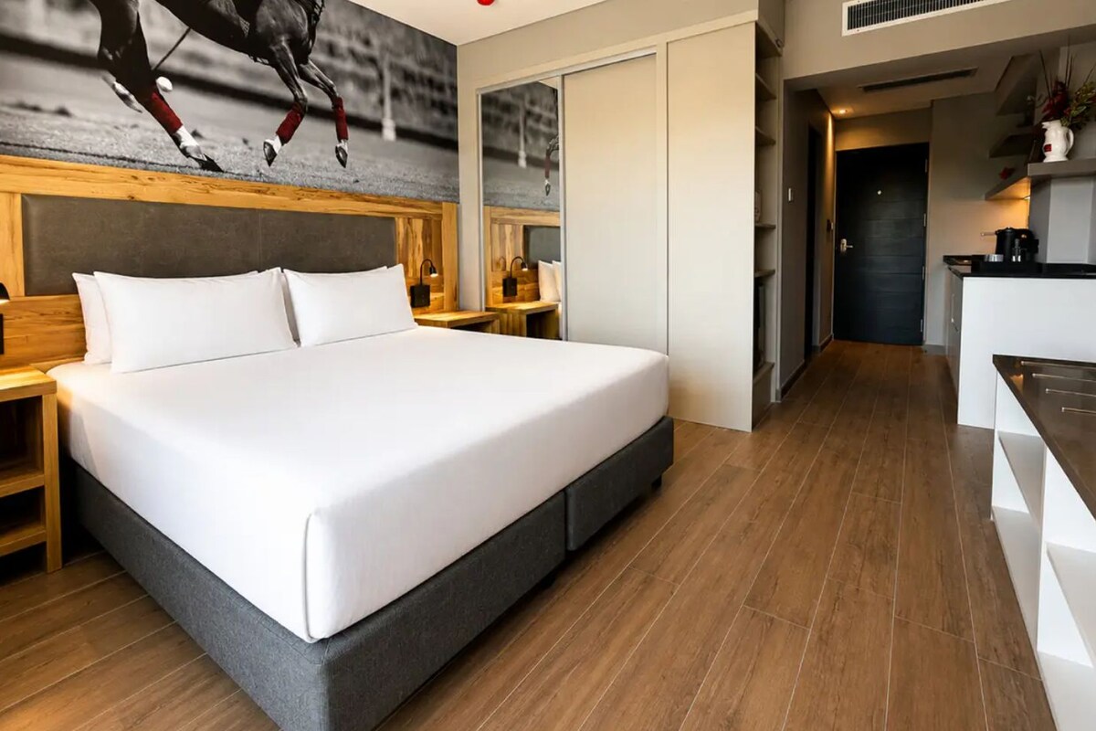 A modern hotel room with a large bed, wooden headboard, and a kitchen area.