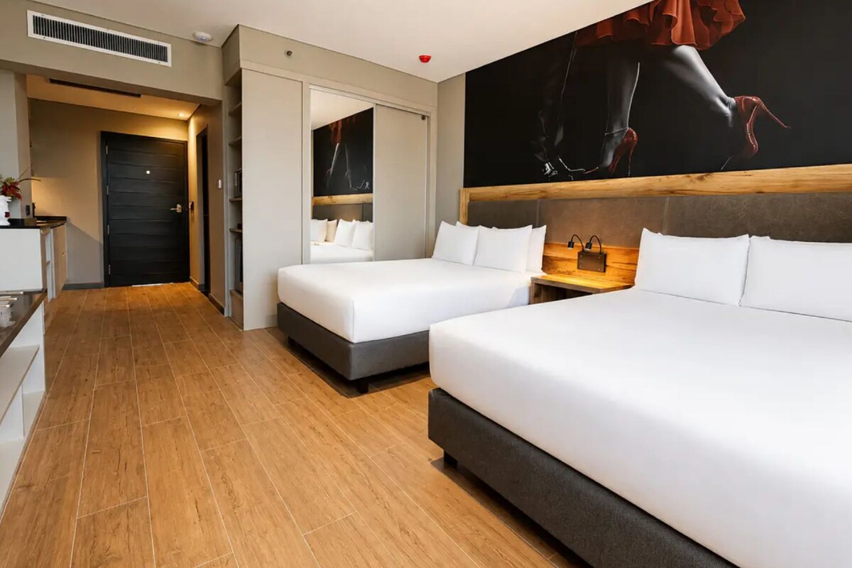 A hotel room with two beds, a wooden floor, a large painting on the wall, and a small table with a lamp.