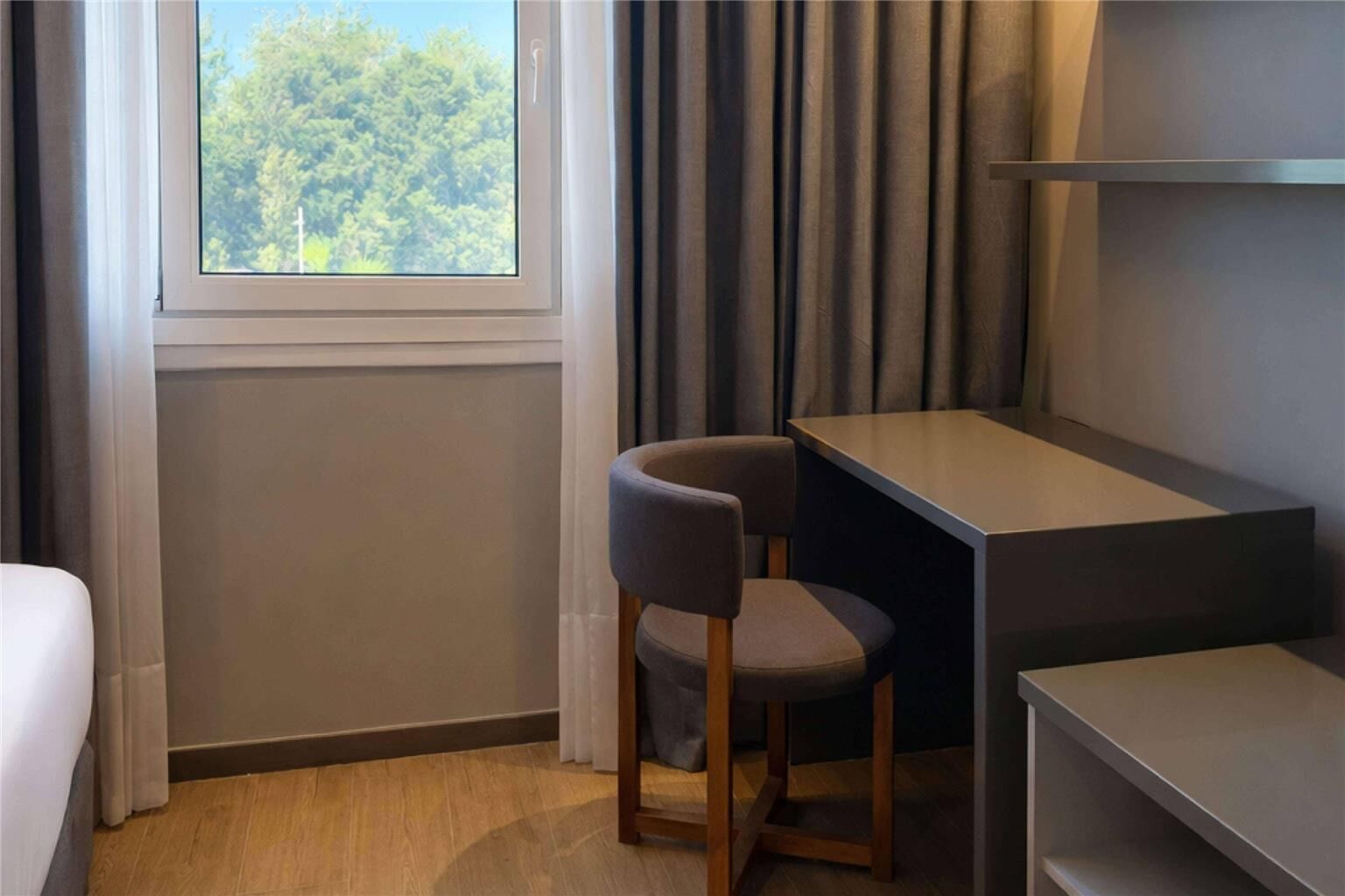 A hotel room with a desk, chair, and a window with a view of trees.