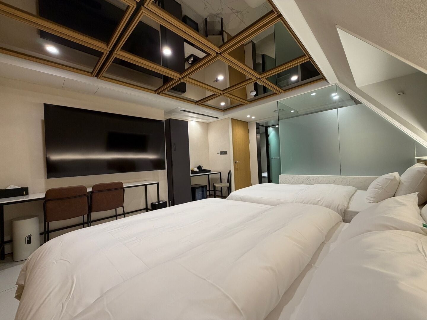 A modern hotel room with a large bed, a desk with chairs, a TV, and a glass-enclosed balcony.