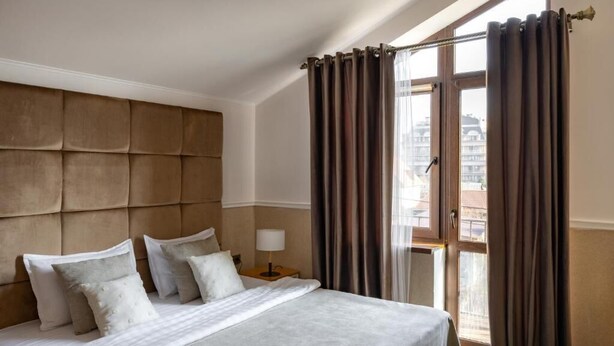 A hotel room with a large bed, a headboard, a nightstand with a lamp, and a view of the city through a window with curtains. - Mountain View Boutique Almaty (Almaty)