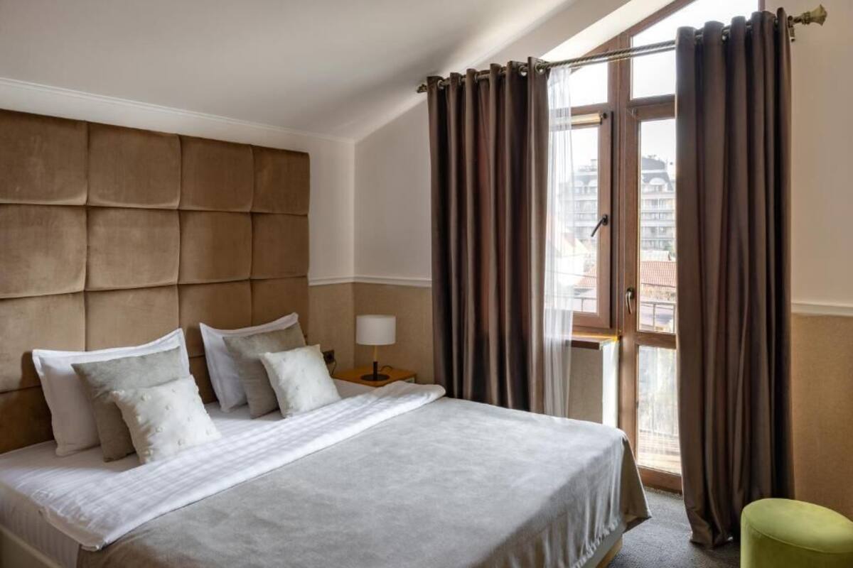 A hotel room with a large bed, a headboard, a nightstand with a lamp, and a view of the city through a window with curtains.