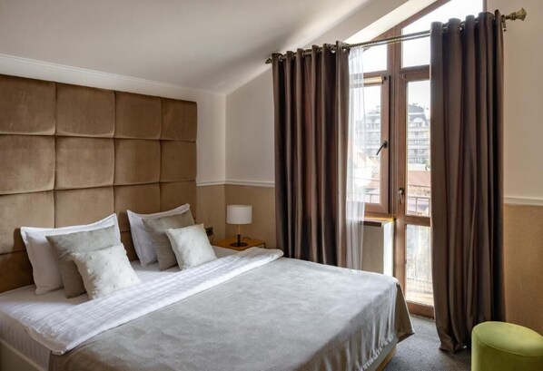 A hotel room with a large bed, a headboard, a nightstand with a lamp, and a view of the city through a window with curtains. - Mountain View Boutique Almaty (Almaty)