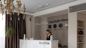 A reception desk with a receptionist, a chandelier, a potted plant, a sign with city names, and a clock. - Mountain View Boutique Almaty (Almaty)