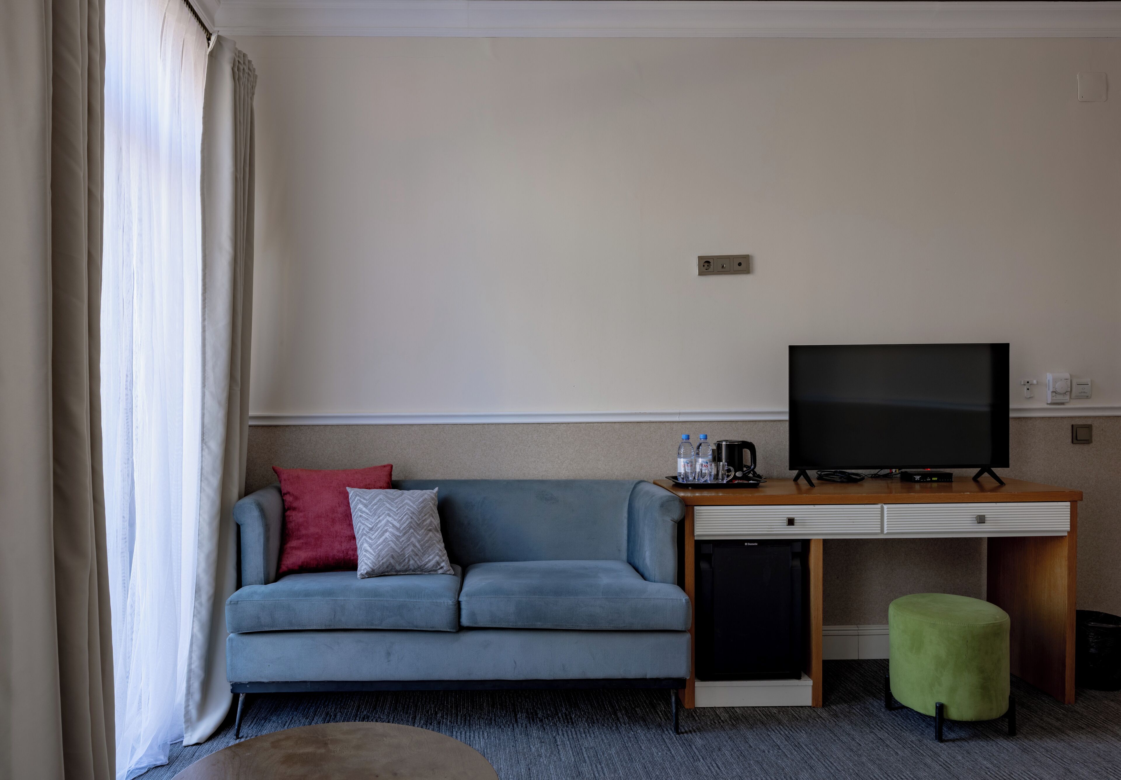 Een hotelkamer met een bank, een televisie, een bureau en een klein, groen voetenbankje.