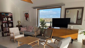 Uma sala de estar moderna com sofá, cadeiras, mesa de centro, televisão, estante e uma janela grande com vista.