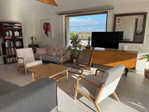 Ein modernes Wohnzimmer mit Sofa, Sesseln, Couchtisch, Fernseher, Bücherregal und einem großen Fenster mit Blick.
