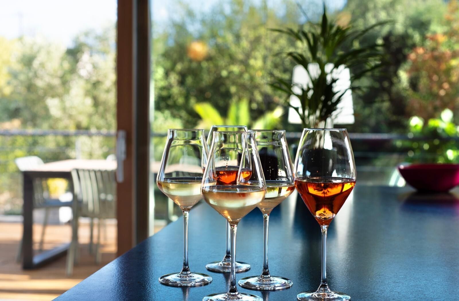 A row of wine glasses on a table with a view of greenery outside.