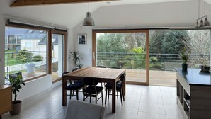 Uma área de refeições moderna com mesa e cadeiras de madeira, janelas amplas e uma varanda com vista para o verde.