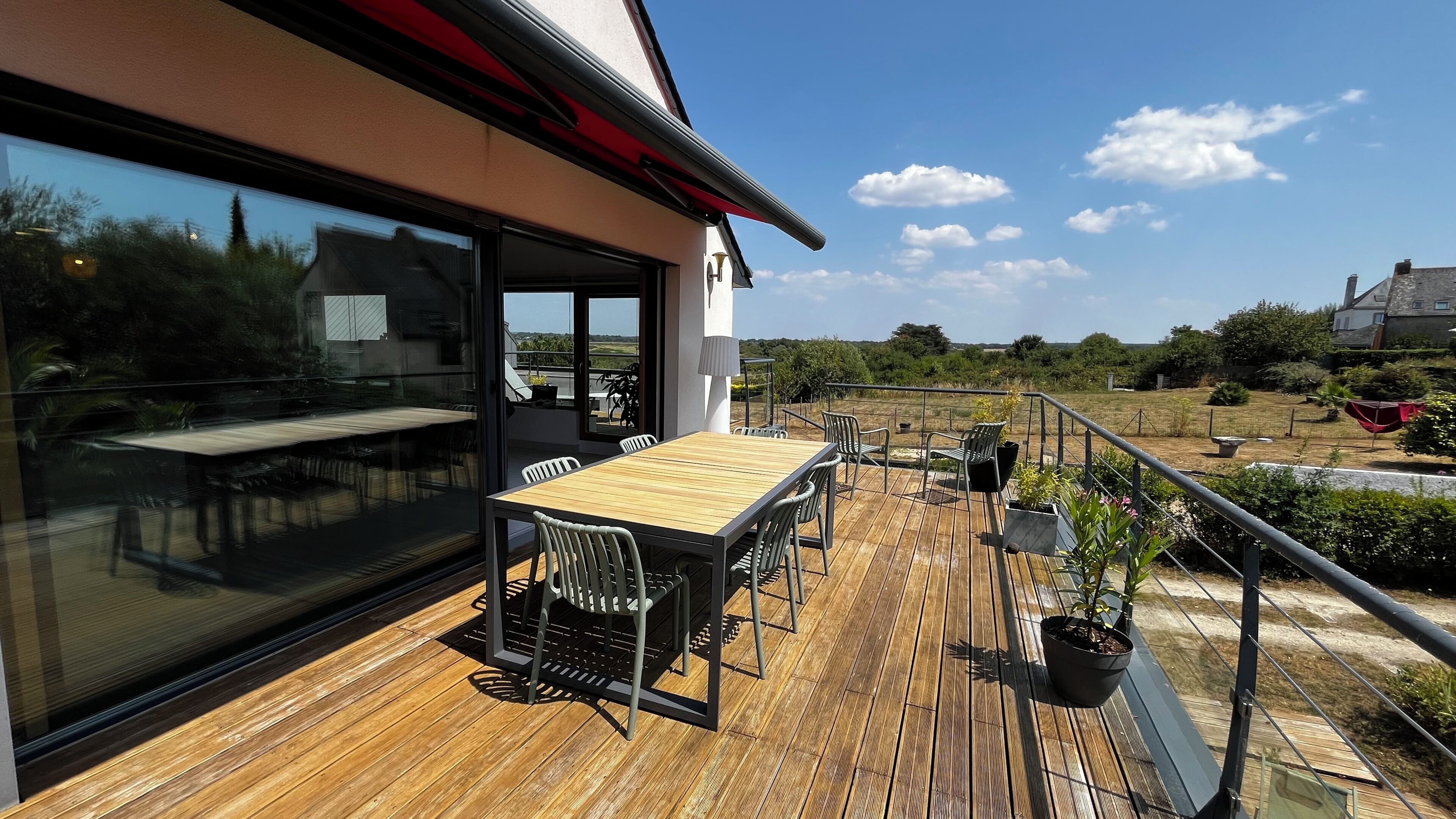 Eine Holzterrasse mit Tisch und Stühlen, eine Glastür, die zu einer Terrasse führt, und ein Blick auf die umliegende Landschaft.