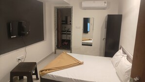 A small, neatly arranged bedroom with a bed, a television mounted on the wall, an air conditioning unit, and a small stool. - Hotel Maruti Inn (Sumerpur)