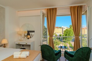 Ein Hotelzimmer mit einem Bett, einem Schreibtisch mit Lampe, zwei Stühlen und einem Balkon mit Blick auf die Stadt.