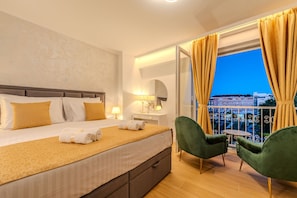 Ein Hotelzimmer mit einem Bett, einem Nachttisch, einer Lampe, einem grünen Sessel, einem kleinen Tisch und einem Balkon mit Vorhängen.