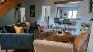 A modern living room with a grey sofa, a white armchair, and a wooden coffee table. There is a staircase in the background, and a kitchen area is visible through an open door. - THE CHARM OF LES BERCEAUX: 5 minutes from Avenue de Champagne and right in the heart of the city (Épernay)