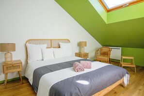 Une chambre avec un plafond en pente, un lit recouvert d’une couette grise et blanche, deux lampes de chevet, une chaise en osier et un petit tabouret en bois.