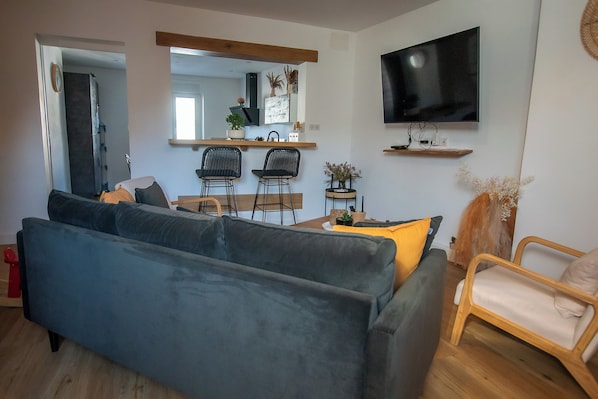 Un salon moderne avec un canapé gris, une table à manger en bois avec des chaises, un téléviseur à écran plat fixé au mur et une petite plante en pot posée sur la table.