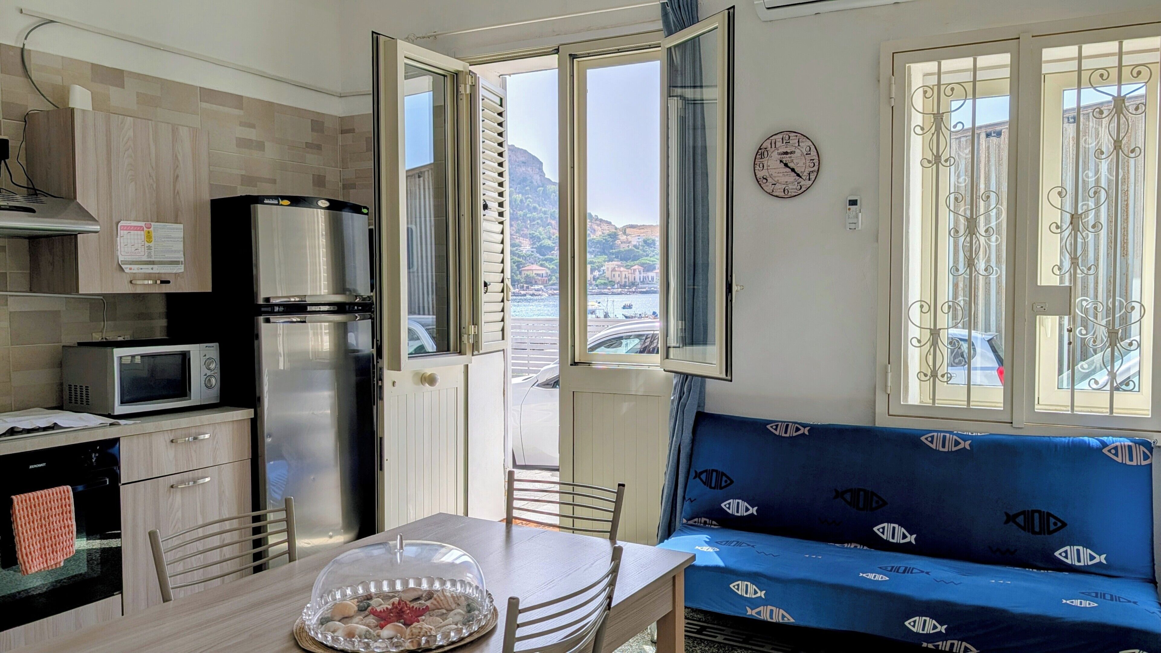 Una kitchenette con microondas, refrigerador y estufa. Hay una mesa de comedor con sillas y un sofá azul con vista al mar.
